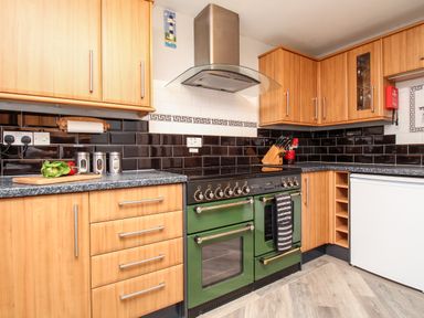 A kitchen with wooden cabinets a green stove a cutting board with vegetables and black tiled backsplash at Knapp Cottage in Chickerell