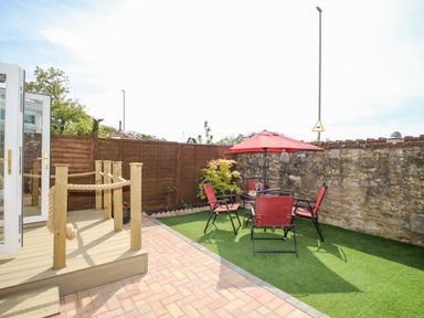 A small outdoor patio with a table and four chairs under a red umbrella next to a wooden deck and stone wall at Knapp Cottage in Chickerell