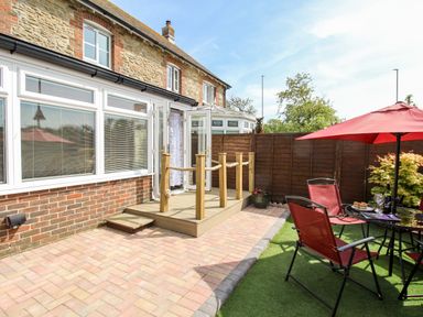A fenced backyard with patio doors leading to a house a small deck a table with red chairs and an umbrella at Knapp Cottage in Chickerell