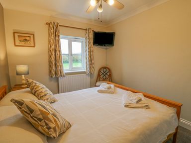 A bedroom with a bed pillows folded towels a chair a window with curtains a wall-mounted television and a bedside lamp at Knapp Cottage in Chickerell