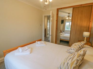 A bedroom with a bed with white linens and patterned pillows a wooden wardrobe with a mirror and an open door showing a window at Knapp Cottage in Chickerell
