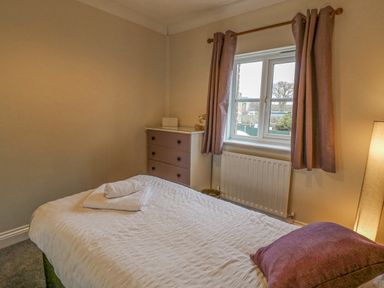 A bedroom with a bed covered in white bedding and a purple pillow a chest of drawers a window with curtains and a floor lamp at Knapp Cottage in Chickerell