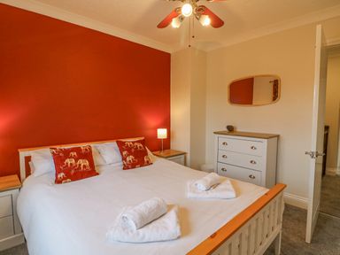 A bedroom with a double bed white bedding red pillows two bedside tables a dresser with a mirror and an open door at Knapp Cottage in Chickerell