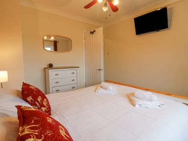 A bedroom with a bed with red pillows white towels a chest of drawers a wall-mounted television and a ceiling fan at Knapp Cottage in Chickerell