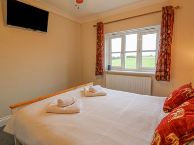 A bedroom with a bed with towels on it red cushions a window with patterned curtains and a wall mounted TV at Knapp Cottage in Chickerell