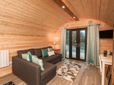 A living room with a dark sofa and cushions a small table with a lamp a geometric rug and glass doors with curtains at Jenny - Crossgate Luxury Glamping in Hartsop near Glenridding