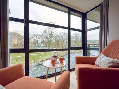 A seating area with two orange chairs and a small round table with a coffee press and two mugs by large windows overlooking a river at Riverside Park 6 in Keswick