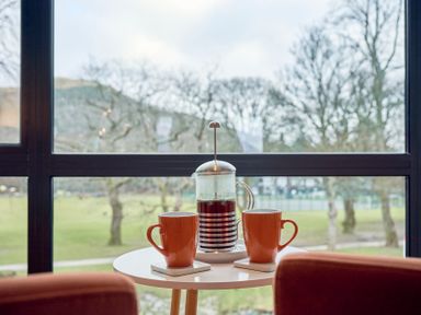 A small round table with two orange mugs and a French press filled with coffee in front of a large window overlooking trees and a park at Riverside Park 6 in Keswick