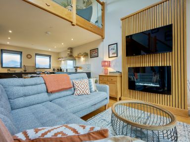A living room with a gray sofa a glass coffee table a wooden sideboard with a lamp a wall-mounted TV and fireplace and kitchen in the background at Riverside Park 6 in Keswick