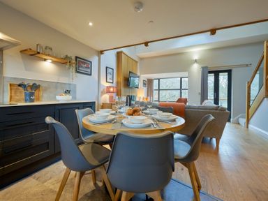 A dining table set with dishes and glasses near a kitchen with cabinets and shelves next to a living area with a sofa and TV at Riverside Park 6 in Keswick