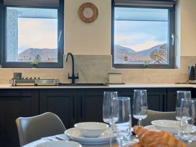 A kitchen with a sink under two windows showing a mountain view with a dining table set with glassware plates and a croissant at Riverside Park 6 in Keswick