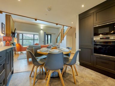 A dining area with a round table and six chairs set with dishes and glasses next to a kitchen with dark cabinets and built-in appliances at Riverside Park 6 in Keswick