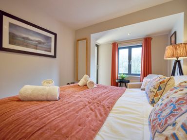 A bedroom with a bed covered with a rust colored blanket and rolled white towels with a window with orange curtains at Riverside Park 6 in Keswick