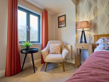 A bedroom corner with a beige armchair with patterned cushion a black round side table with a plant red curtains and a wooden bedside table with a lamp at Riverside Park 6 in Keswick