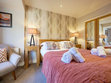 A bedroom with a bed with patterned pillows and folded towels bedside tables with lamps an armchair and a mirrored wardrobe at Riverside Park 6 in Keswick