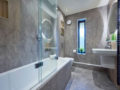 A bathroom with a bathtub a wall-mounted sink a toilet a tall narrow window and a round illuminated mirror at Riverside Park 6 in Keswick