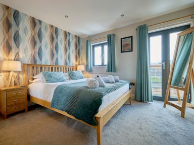 A bedroom with a wooden bed and bedside table with lamps blue bedding and curtains a standing mirror and a balcony door at Riverside Park 6 in Keswick