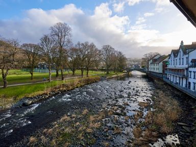 A river flowing between trees and buildings with a walking path on one side at Riverside Park 6 in Keswick