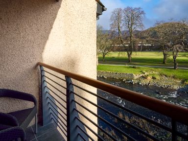 A balcony with two chairs overlooking a river and a grassy park with trees at Riverside Park 6 in Keswick