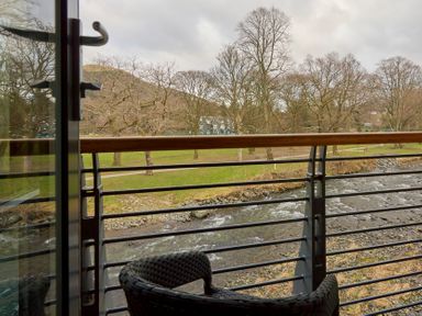 A balcony with a chair overlooking a river and park with trees at Riverside Park 6 in Keswick