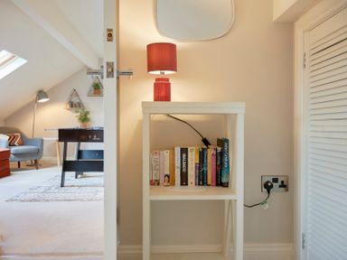 A small white table with a red lamp and books next to a door and a room with a sofa and foosball table at Riverside Park 6 in Keswick