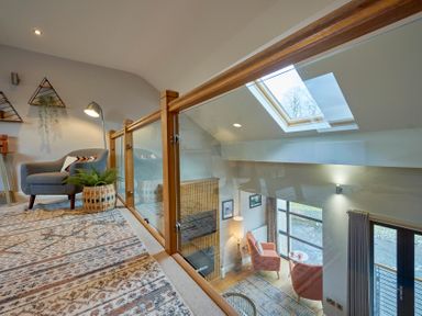 A sitting area with a gray chair, floor lamp, plant in basket, and triangular wall shelves on upper level overlooking a living room with pink chairs and large windows at Riverside Park 6 in Keswick