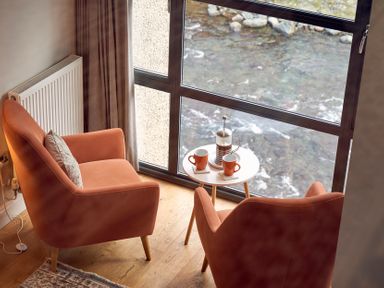 Two orange armchairs with a small round table holding two cups and a coffee press by a large window overlooking a river at Riverside Park 6 in Keswick