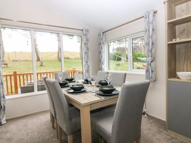 A dining room with a wooden table set for six with gray chairs and large windows showing an outside view at Woodpecker Lodge in Dunbar