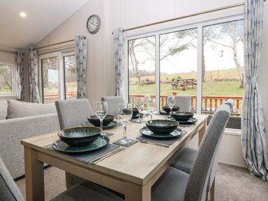 A dining area with a table set for six beside large windows overlooking a grassy outdoor area at Woodpecker Lodge in Dunbar