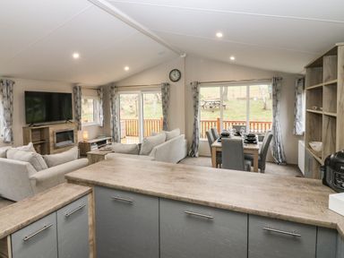 An open living space with a kitchen counter in the foreground sofas and a television on a stand in the living area dining table with chairs set for meals and large windows with curtains at Woodpecker Lodge in Dunbar