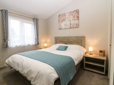 A bedroom with a double bed with white and blue bedding two bedside tables with lamps and a window with grey curtains at Woodpecker Lodge in Dunbar