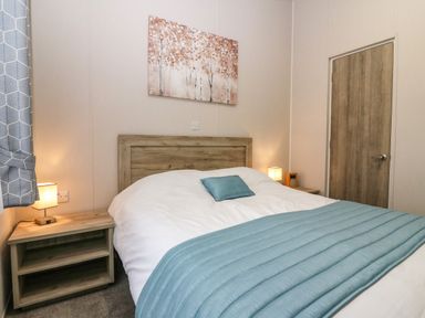 A bedroom with a wooden bed frame a white duvet blue blanket and pillow lamps on wooden bedside tables and a painting of trees on the wall at Woodpecker Lodge in Dunbar