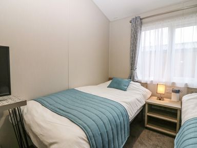 A bedroom with two single beds blue bed runners a bedside table with a lamp and window with curtains at Woodpecker Lodge in Dunbar