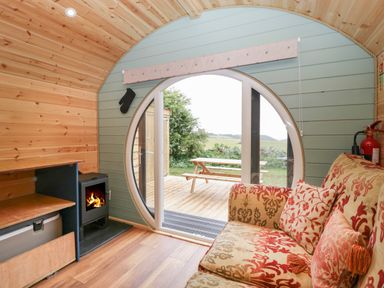 A small room with a patterned sofa a wood-burning stove and a round window looking out to a picnic table and greenery at Lime Kiln in Blagdon