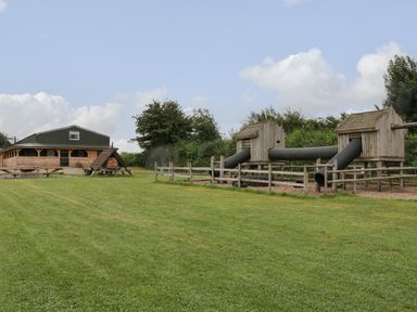An outdoor play area with two wooden playhouses connected by tunnels and a wooden building in the background at Lime Kiln in Blagdon