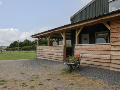 A wooden structure with a covered porch and a wheelbarrow with flowers outside Lime Kiln in Blagdon