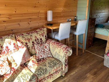 A wooden interior room with a patterned sofa a tall wooden table with two chairs and a bed visible through a door at Lime Kiln in Blagdon