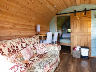 An interior view of a wooden room with a patterned sofa, a table with two white chairs, and a sliding wooden door leading to a bedroom at Lime Kiln in Blagdon
