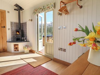 An interior with a wood stove, open door leading outside, wall-mounted clothes hangers, and flowers on a wooden surface at Balwen Shepherd's Hut in Berriew