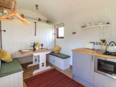 A small kitchen with a wooden table and green cushioned benches a countertop with an oven kettle and toaster and a wall shelf with cups at Balwen Shepherd's Hut Berriew