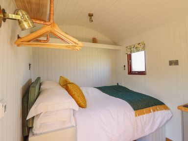 A small bedroom with a bed with white bedding and green and yellow cushions and a window with floral blinds at Balwen Shepherd's Hut in Berriew