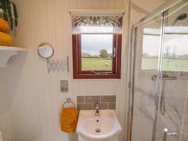 A bathroom with a sink, shower area, window with a roller blind, round extendable mirror, and yellow towels at Balwen Shepherd's Hut in Berriew