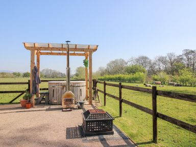 An outdoor area with a wooden hot tub under a pergola and a fire pit near a fenced grassy field with sheep at Balwen Shepherd's Hut in Berriew