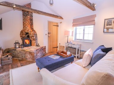 A living room with a brick fireplace wood stove beige sofa blue ottoman wooden door and window at The Forge in Old Newton near Stowmarket
