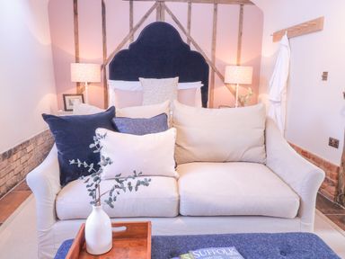 A bedroom with a beige sofa with cushions in front of a bed with a dark headboard and bedside lamps at The Forge in Old Newton near Stowmarket