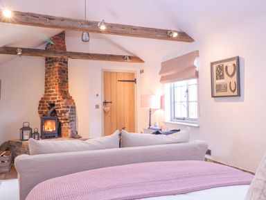 A living room with a brick fireplace wooden beams a beige sofa a wooden door and a window with a lamp at The Forge in Old Newton near Stowmarket