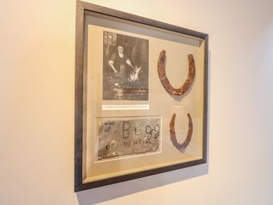 A framed black and white photo of a blacksmith with two horseshoes and a metal plaque signed J Bugg Old Newton at The Forge Old Newton near Stowmarket