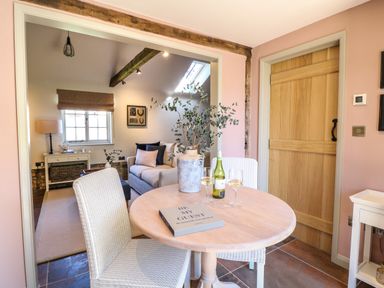 A dining area with a round wooden table, two chairs, a book, a vase with branches, two wine glasses, and a bottle of wine next to a wooden door and a living room with a couch and a window at The Forge in Old Newton near Stowmarket