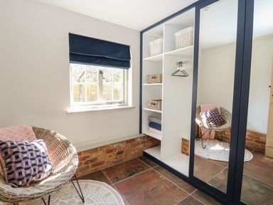 A small room with a wicker chair with cushions a window with a navy blind a white shelving unit with baskets and a mirrored wardrobe door at The Forge in Old Newton near Stowmarket