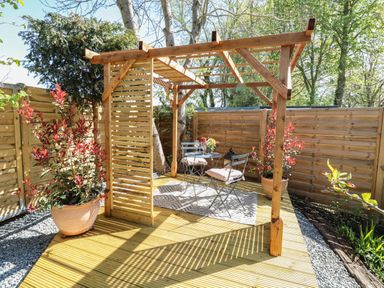 A wooden pergola with a small table and two chairs on a deck surrounded by plants at The Forge in Old Newton near Stowmarket